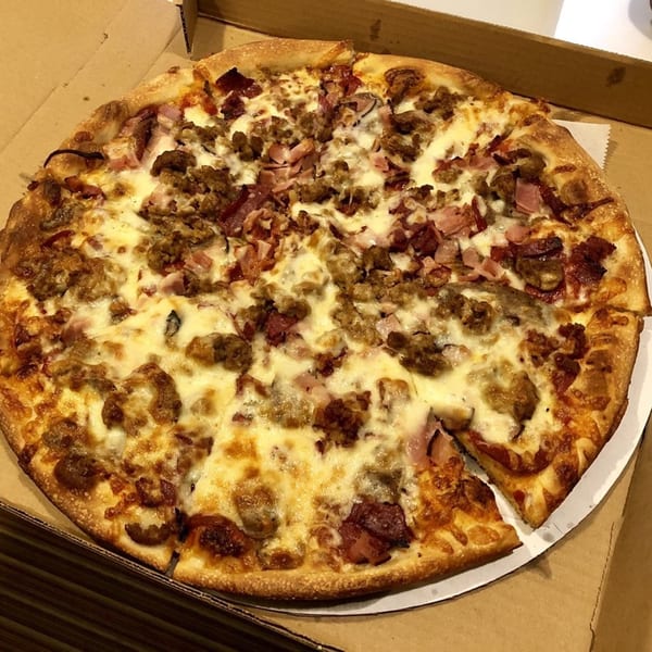 A whole Anastasia's Pizza with various meat toppings and melted cheese, in a cardboard box.