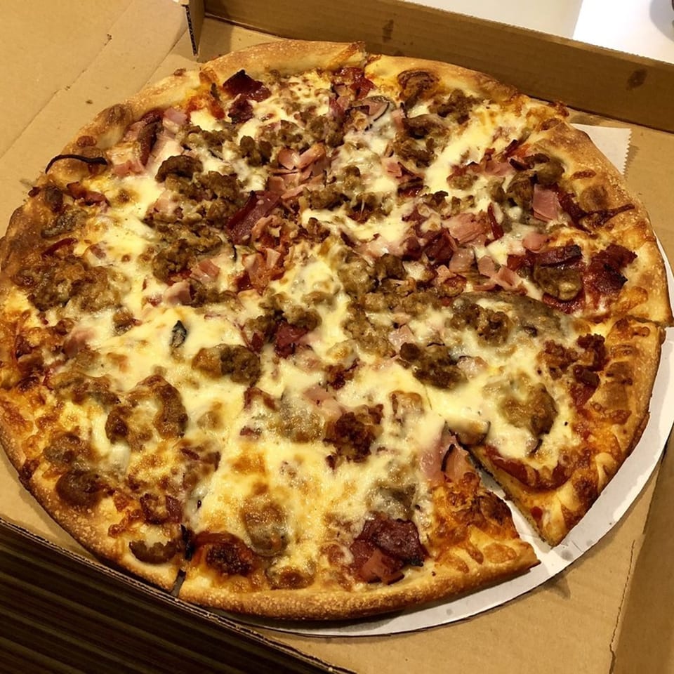 A whole Anastasia's Pizza with various meat toppings and melted cheese, in a cardboard box.