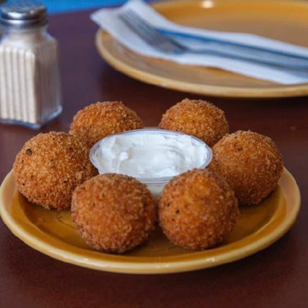 Six golden-brown fried arancini balls arranged around a small cup of white dipping sauce on a yellow plate.